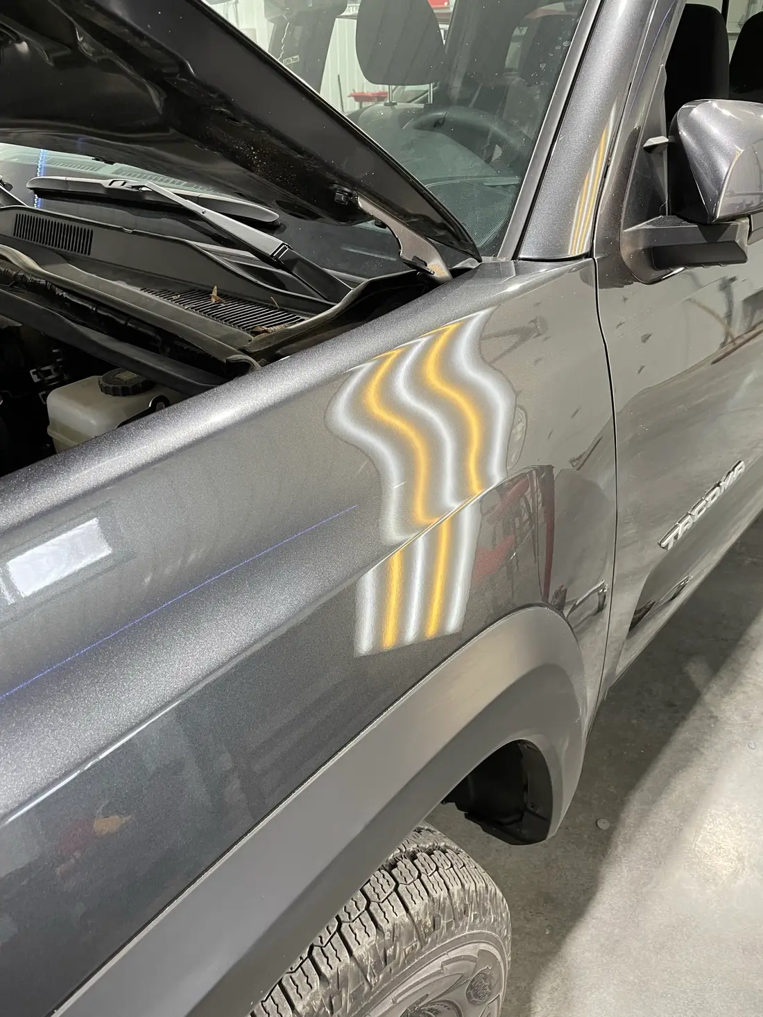 Dark grey Toyota Tacoma fender panel showing hail dents revealed by LED line-board reflection